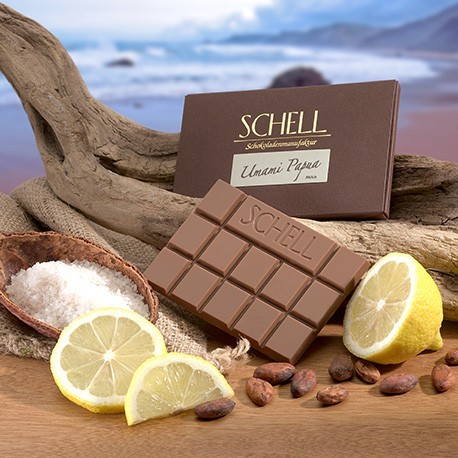 Die Verpackte Umami Papua Schokolade lehnt an ein geschwungenen Holz, darunter eine halbe Zitrone mit der unverpackten Schokolade, einer halben Kakaoschote gefüllt mit Meersalz und Zitronenscheiben. Es ist eine Milchschokolade mit einer feinen Citrusnoten und Meersalz