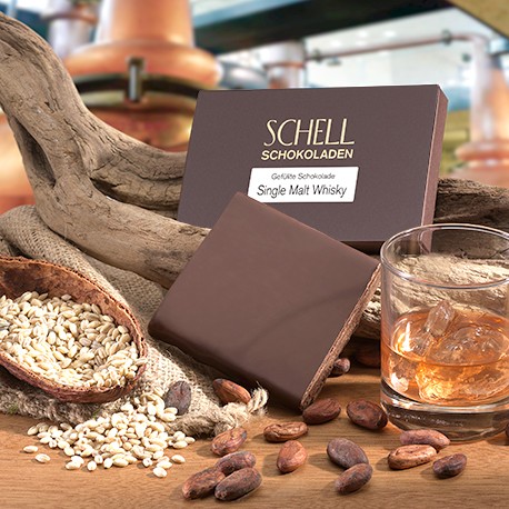 Whisky Single Malt in der Verpackung lehnt an ein geschwungenes Holz, darunter die unverpackte Schokolade und eine halbe Kakaoschote gefüllt mit Nüssen. Rechts von der Schokolade befindet sich ein Glas mit Whisky gefüllt.