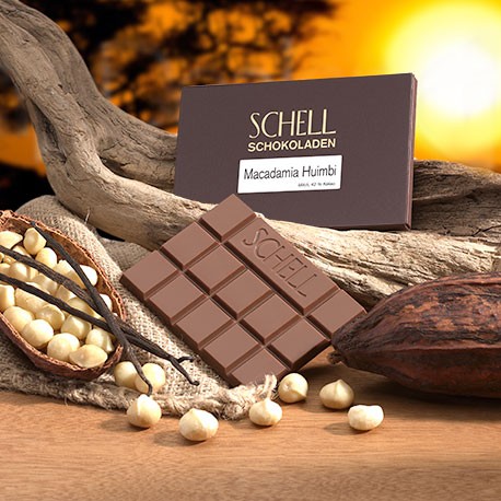 Die verpackte Schokolade lehnt an ein geschwungenes Holz, darunter die nicht verpackte Milchschokolade und eine Kakaoschote gefüllt mit Macadamia Nüssen.
