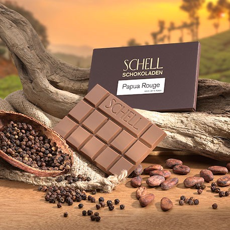Die verpackte Schokolade lehnt an ein geschwungenes Holz, darunter die nicht verpackte Milchschokolade und eine Kakaoschote gefüllt mit rotem Pfeffer.